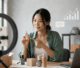 Woman filming a beauty product review with a ring light, makeup bottles on a desk, and a laptop nearby.