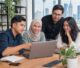 Four colleagues of diverse backgrounds gather around a laptop at a wooden table in a bright office, collaborating with a city skyline outside.